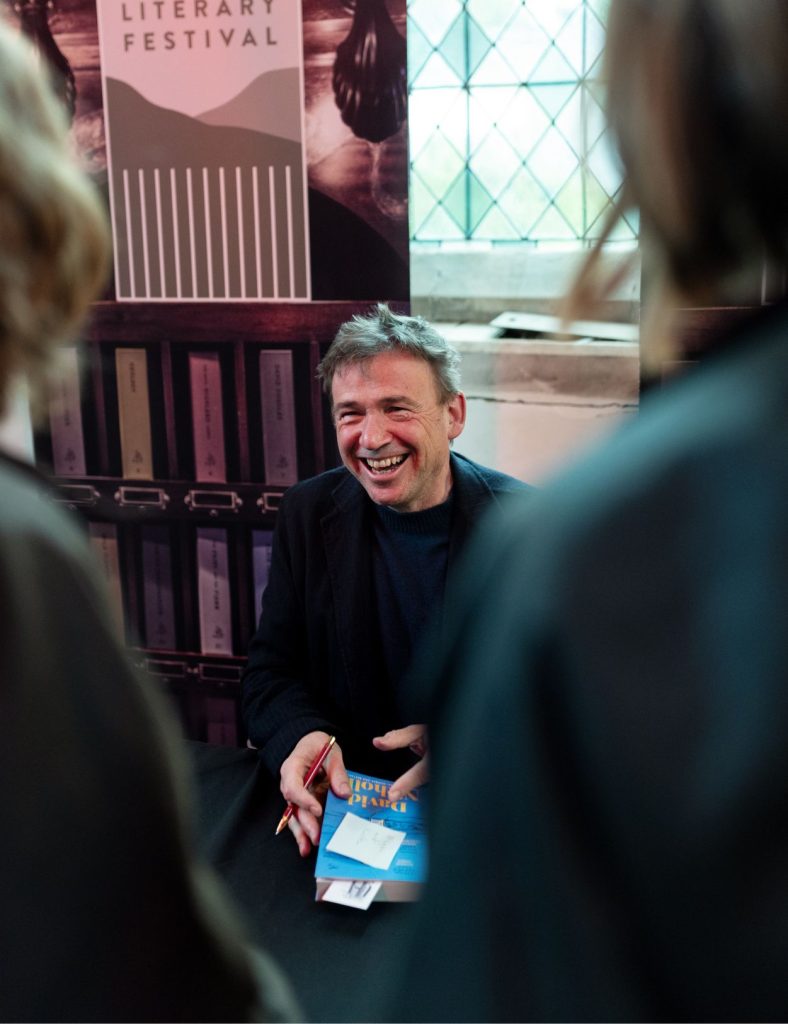 Speaker David Nicholls sitting down smiling, about to sign one of his books with two female guests watching.