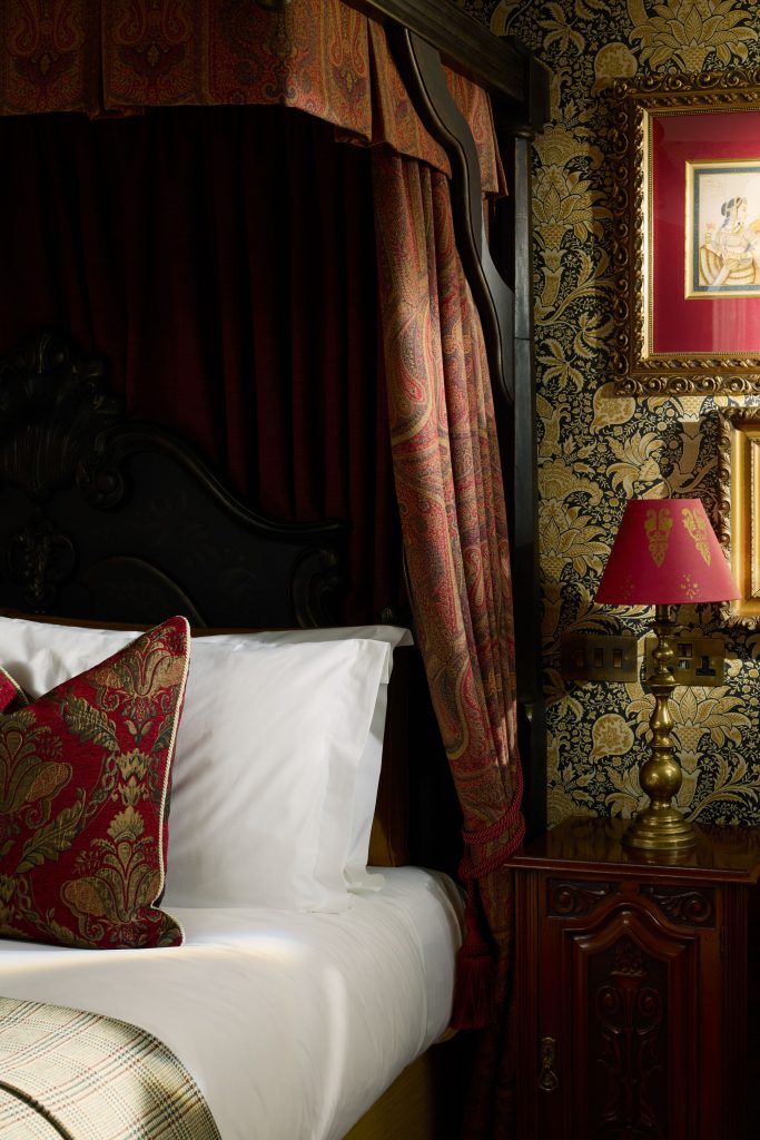 A corner of a bed with a bedside table, lamp and red furnishings