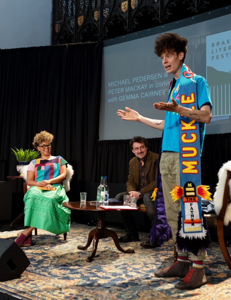Three speakers on stage at the Braemar Literary Festival with one standing up addressing a crowd, and the other two sitting down smiling at him.