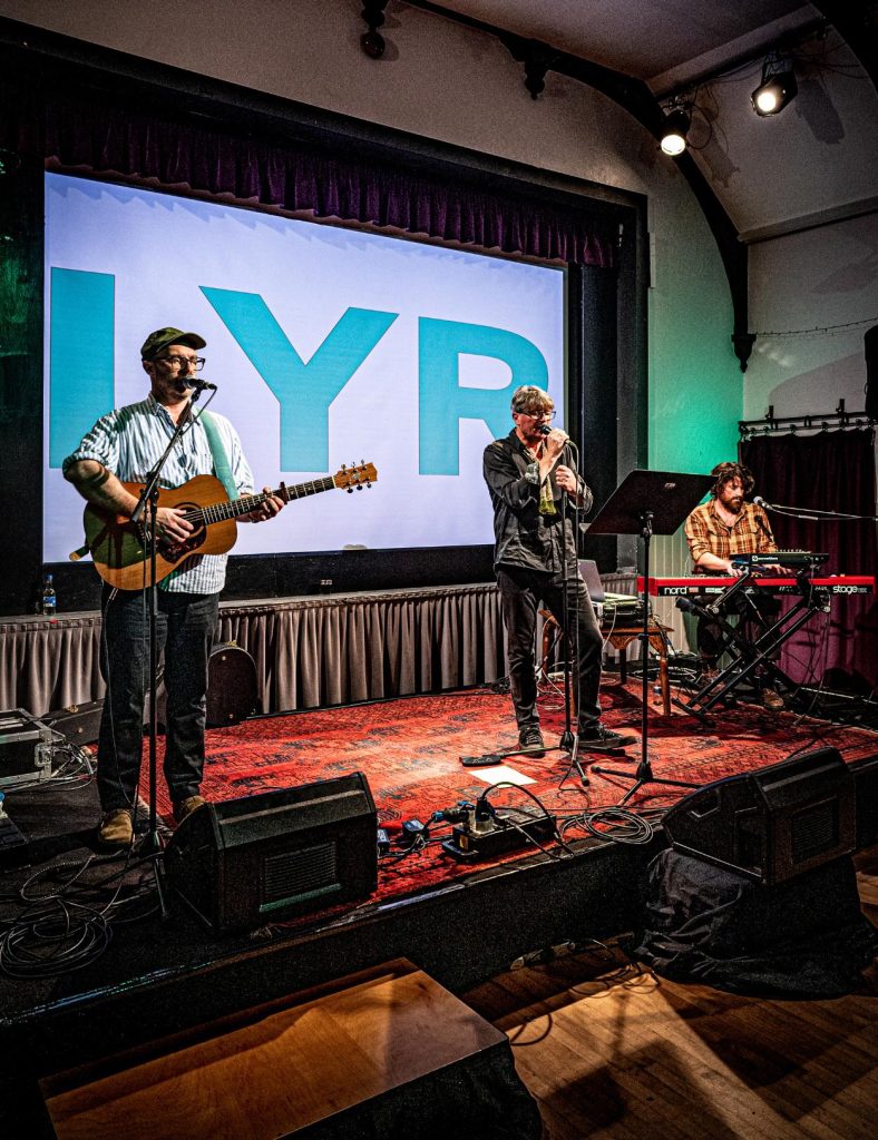 Three musicians in the band 'LYR' sing together on a red carpet stage, with one man playing the guitar and another the electric keyboard.