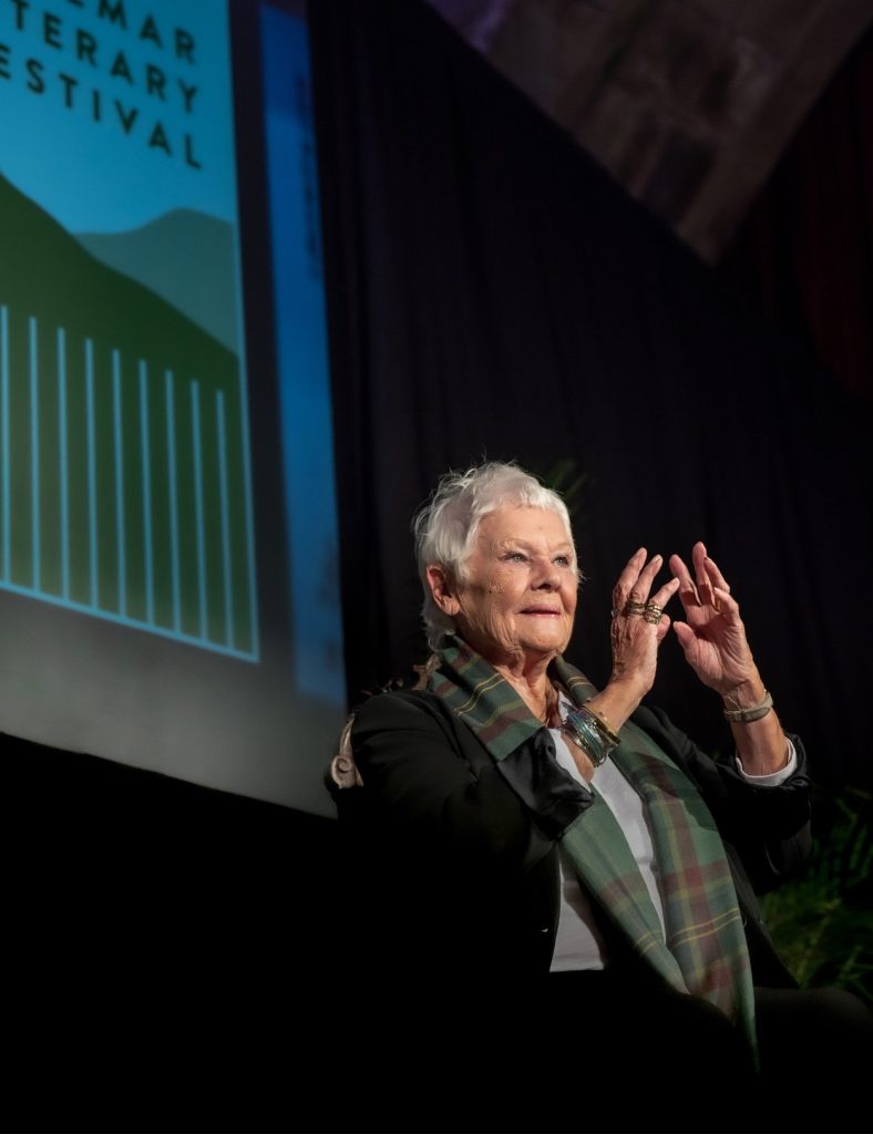 Speaker Dame Judi Dench standing on stage wearing a tartan scarf, addressing a crowd at the Braemar Literary Festival.
