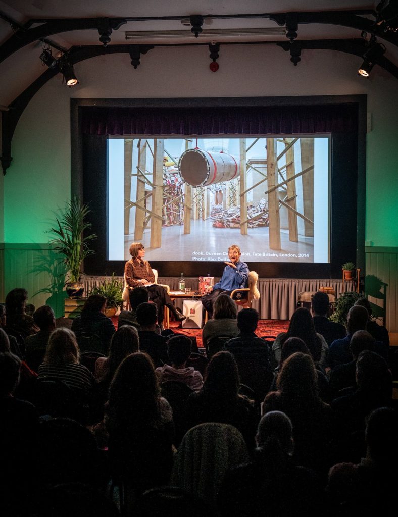 Speakers Frances Morris and Fiona Bradley sit on a red carpet stage overlooking an audience with a big screen in the background.