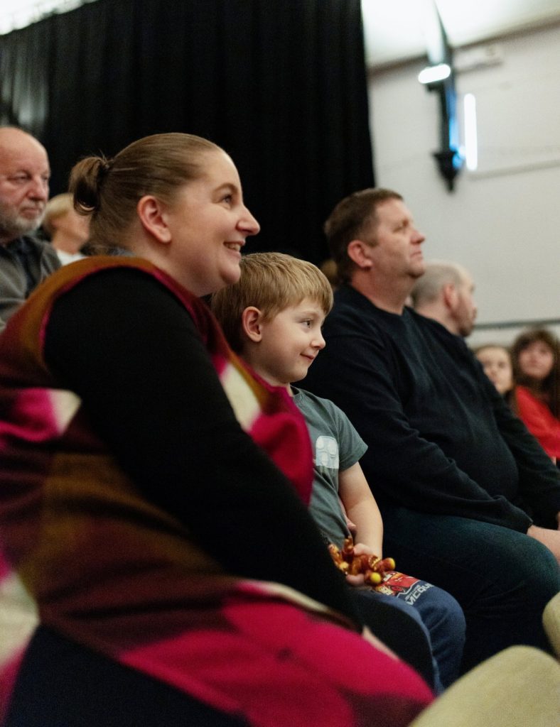 A child sits smiling between his parents in a group of people watching Suzie Edge perform at the Braemar Literary Festival.