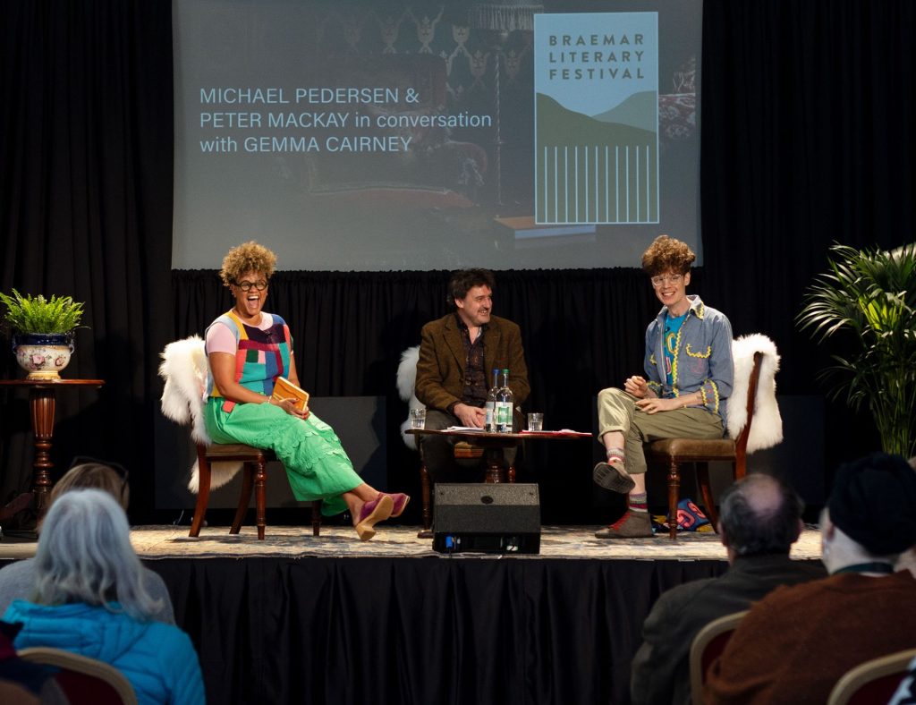 Three speakers sitting on a stage looking out to an audience with a big screen behind them saying 'Michael Pedersen & Peter Mackay in conversation with Gemma Cairney'.
