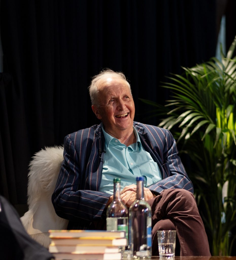 A close-up of speaker Sir Alexander McCall Smith smiling out at a crowd whilst seated on a stage wearing a blue stripe suit and turquoise shirt.