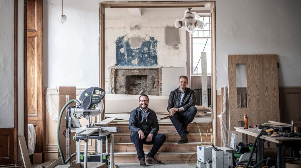 Two brother sit on wooden steps in a partially renovated Flying Stag filled with construction tools and materials, with an exposed fireplace and unfinished walls.