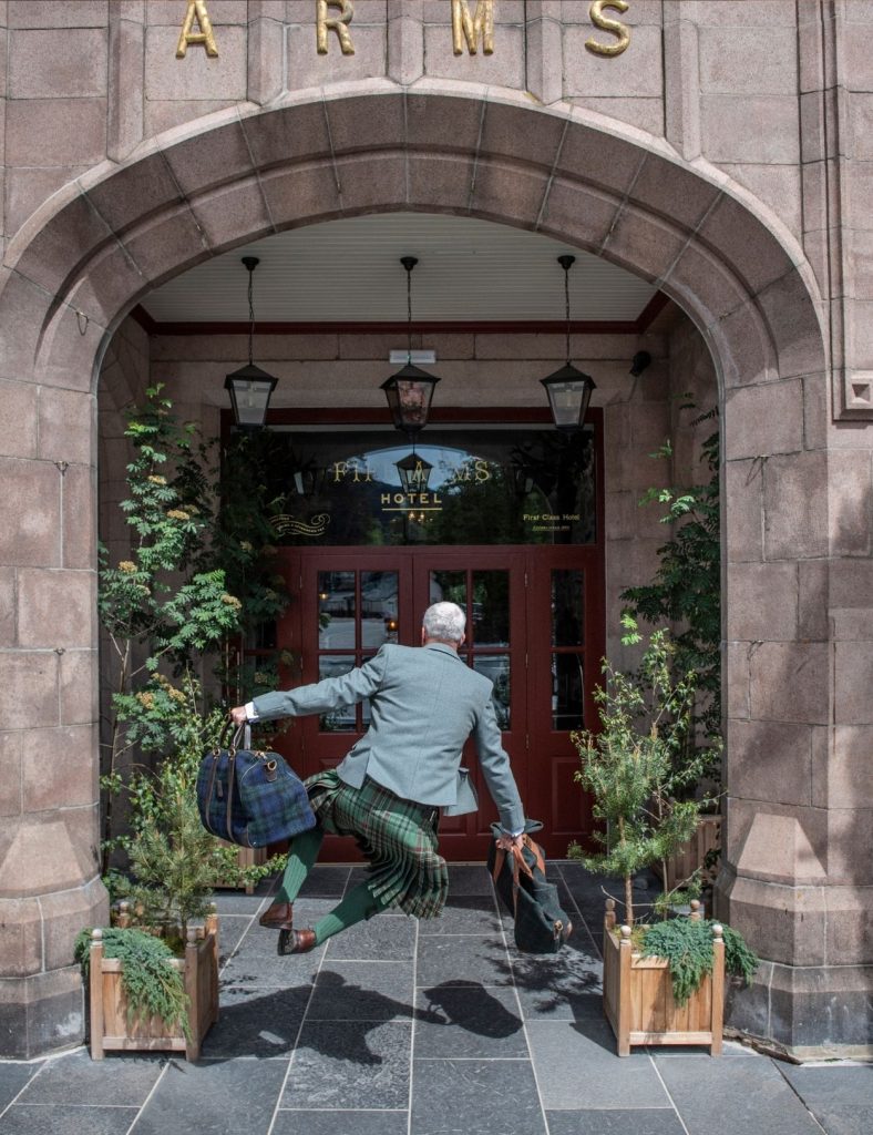 Jumping ghillie dressed in a kilt outside the fife arms hotel main entrance carrying two bags.