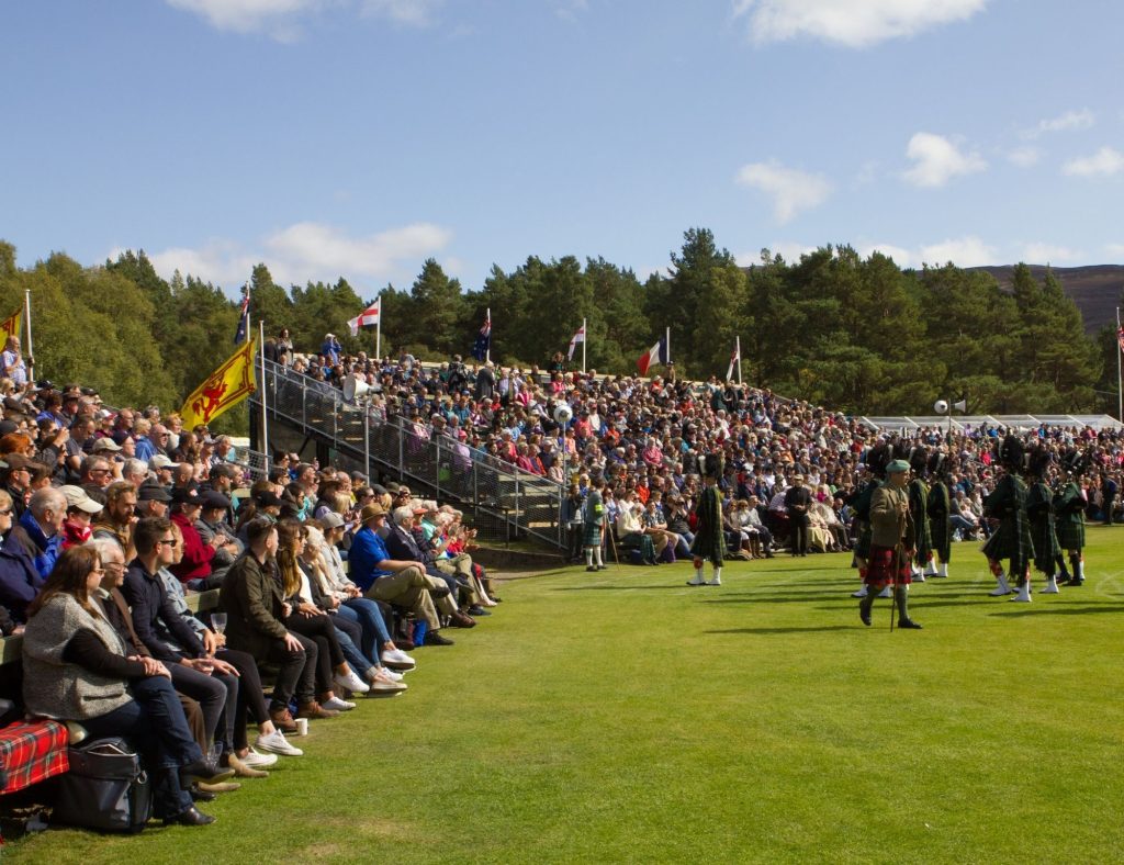 Crowds seated in grandstands watch kilted performers march across a sunny field at an outdoor event in the Scottish Highlands.