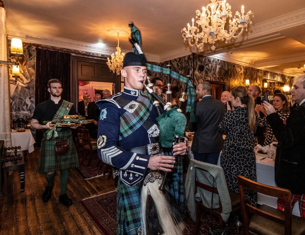 A Scottish piper in kilt uniform is followed by a server carrying a haggis, while guests stand and watch under ornate chandeliers.